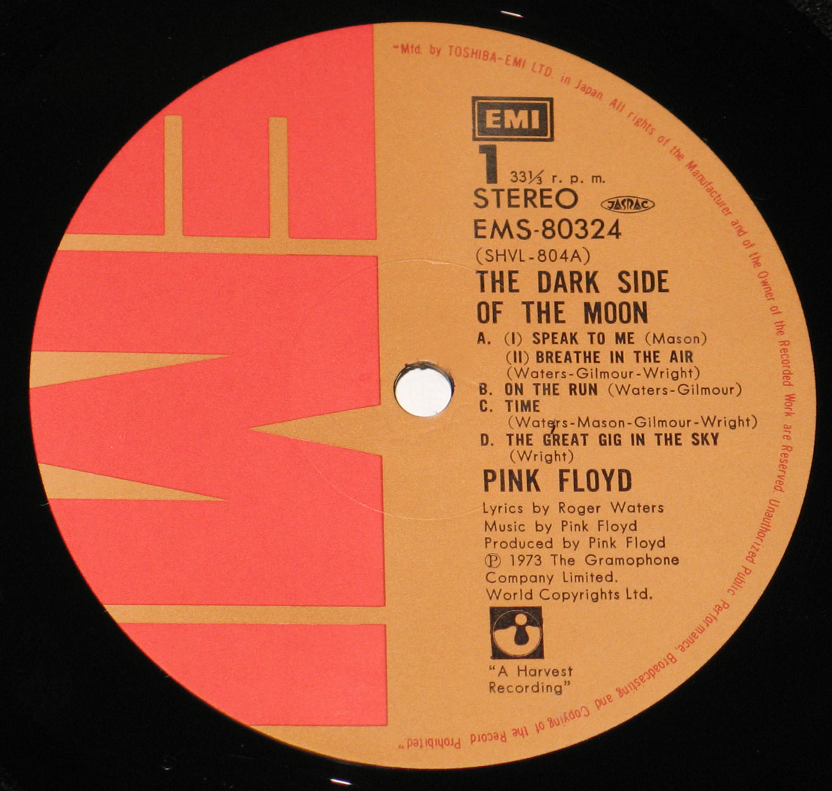 ‘Record label of Pink Floyd’s The Dark Side of the Moon Japanese edition. Features an orange and red background with an EMI logo and catalog number EMS-80324 at the top. The label displays essential details, including “33⅓ r.p.m.” and the “JASRAC” logo. Credits for Roger Waters and Pink Floyd appear, along with copyright and licensing information. The label includes “A Harvest Recording” and the Harvest logo, and the edge text notes “Mfd. by TOSHIBA-EMI LTD. in Japan,” indicating Japanese manufacturing.’